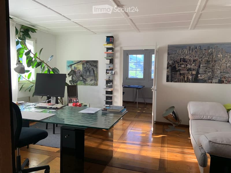 Bright, open room with wooden flooring, a glass desk, a sofa, a chair, a screen, and a lamp. There are books, a plant, and artwork on the walls.