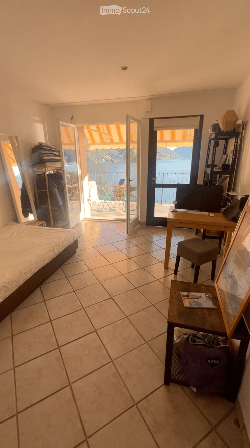 Bright bedroom with tiled floor, bed, desk, and open sliding doors leading to a balcony with a scenic lake view.