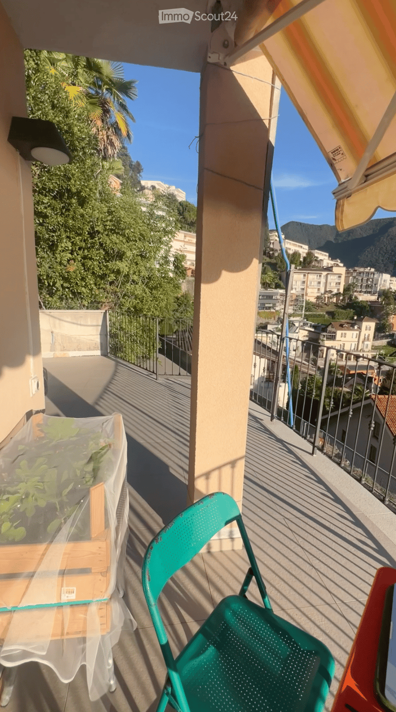 Balcony with a view of the city and mountains, featuring a green plastic chair and a covered table with plants.
