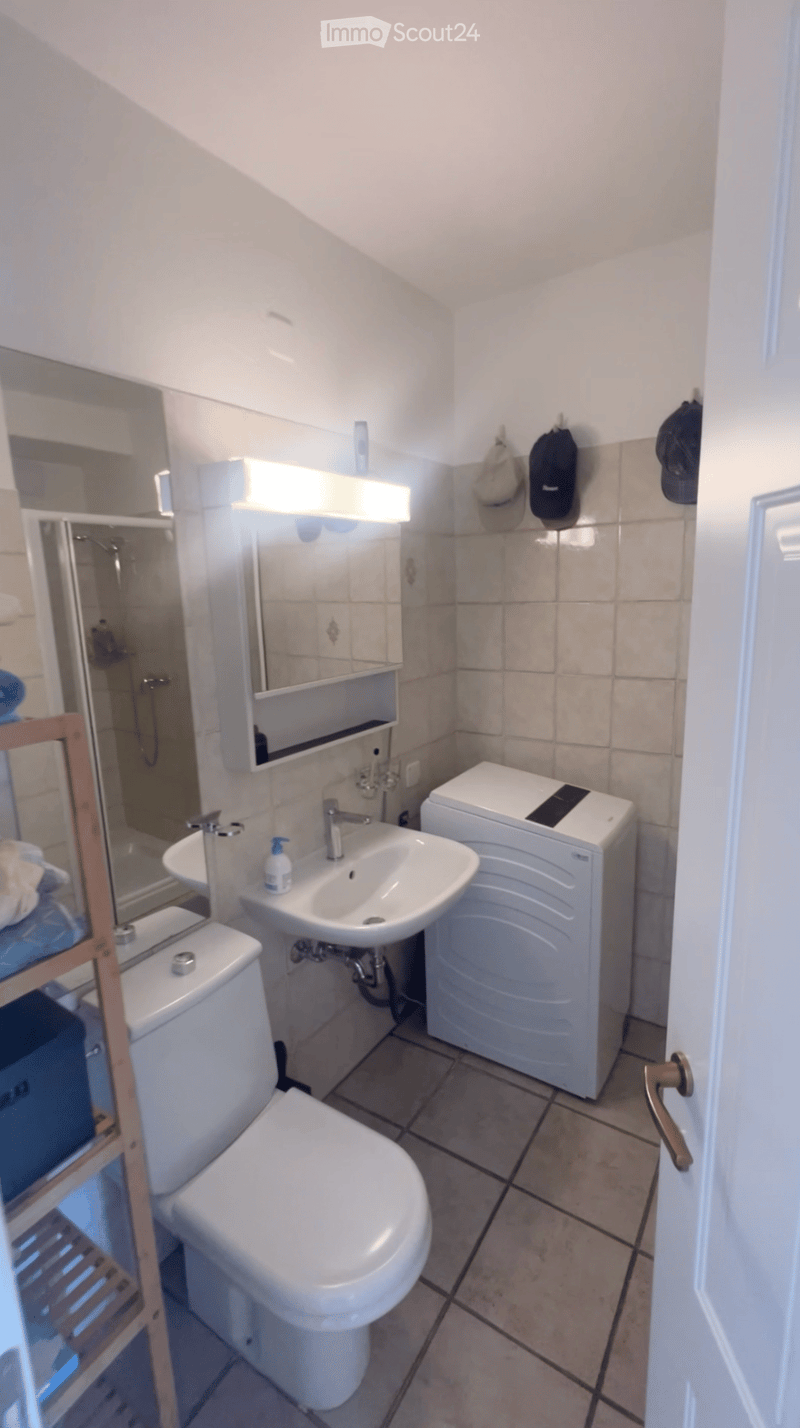 Modern bathroom with white tiles, a toilet, sink, mirror, and a small shower area. Includes a washing machine and a wooden shelf with folded towels.