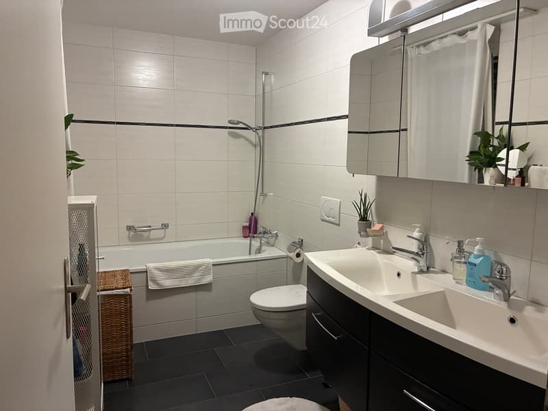 Modern bathroom with white tiles, a bathtub, toilet, double sink, mirror, and various toiletries.