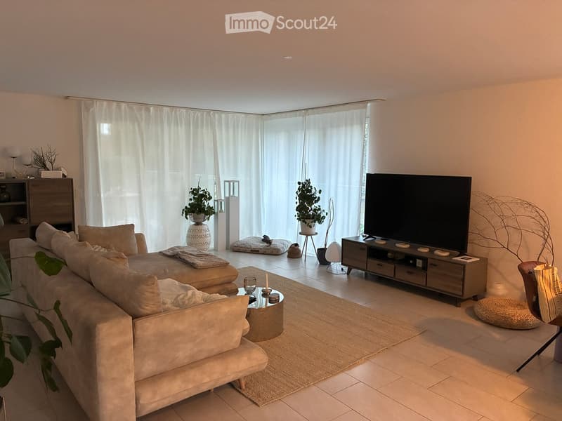 Modern living room with a large sectional sofa, coffee table, TV, side table, and potted plants. The space features large windows with white curtains and a tiled floor.