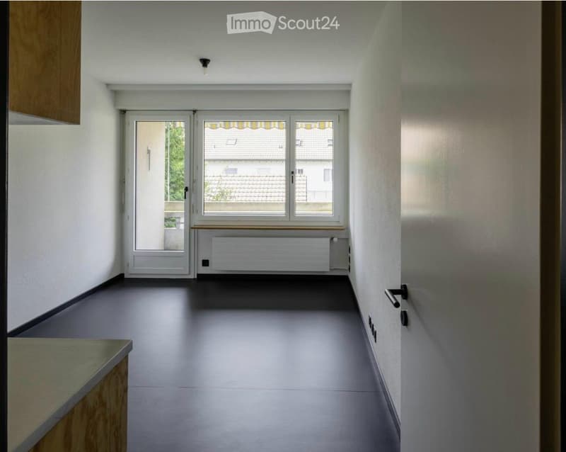 Empty hallway with grey flooring, white walls, a window with blinds, and a radiator below it. The space is clean and unfurnished.
