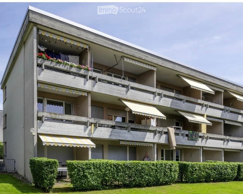 Multi-story apartment building with balconies, awnings, and flower boxes. Green hedges and lawn in front.