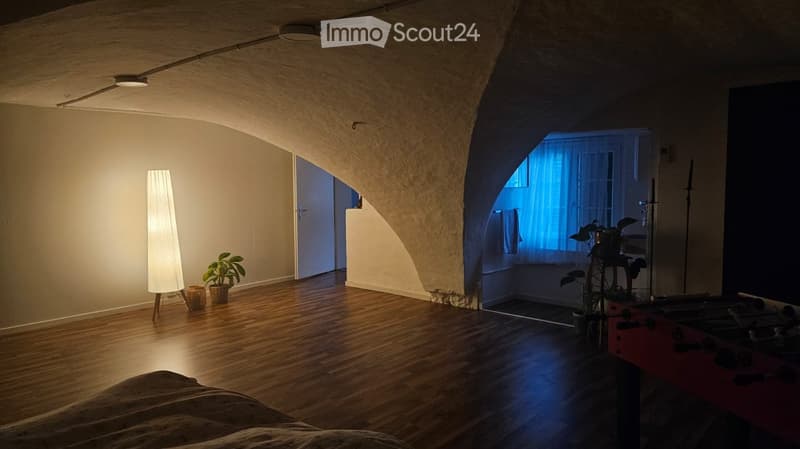 Cozy, well-lit room with wooden flooring, arched doorway leading to a windowed area, a bed, a tall floor lamp, a small plant, and a wicker basket near the entrance.