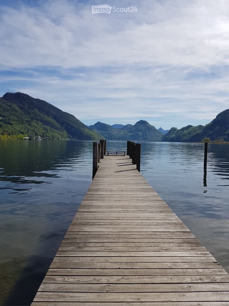 A wooden dock extends into a calm lake surrounded by green, forested mountains under a partly cloudy sky.
