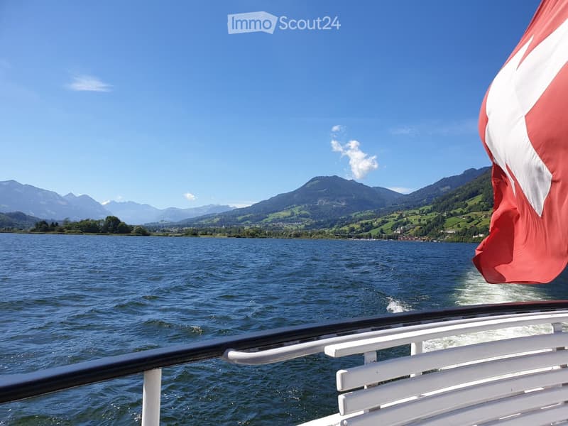 A serene lake view with distant mountains, a Swiss flag visible on the right, and calm water reflecting the clear blue sky.