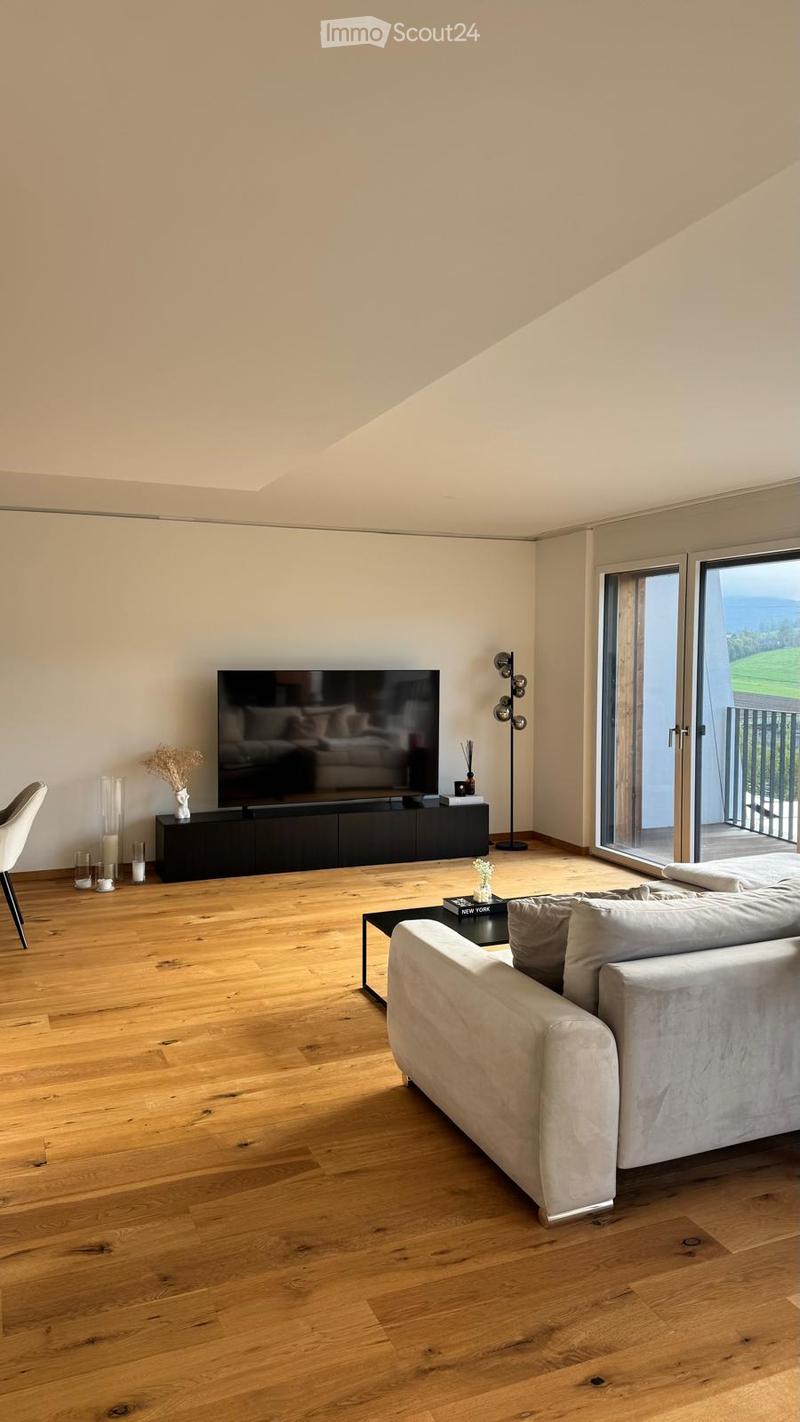 Modern living room with light wooden flooring, large windows offering a scenic view, a grey sofa, coffee table, TV, and decorative items.