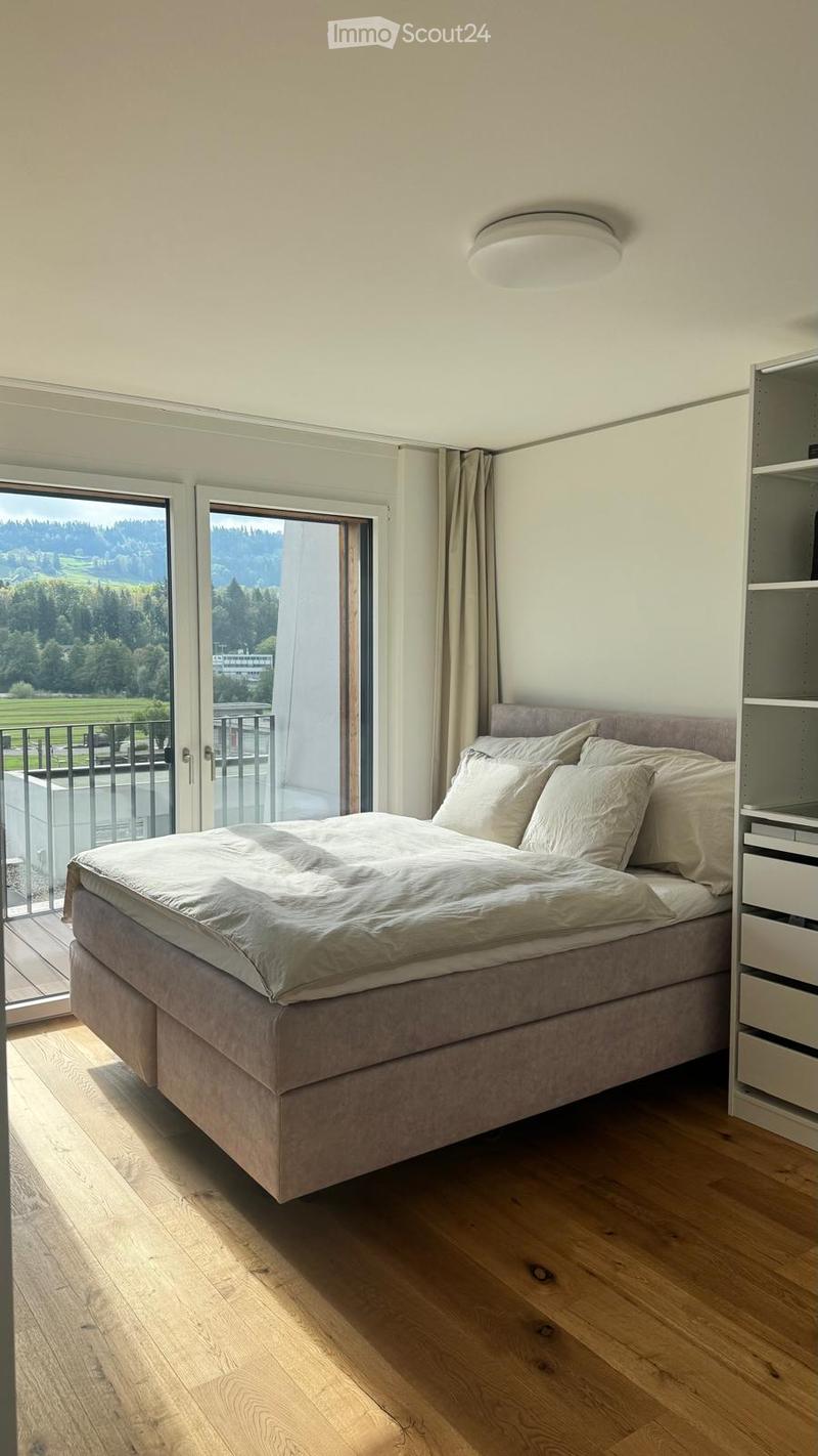 Modern bedroom with a large bed, pillows, nightstand, and a scenic view through large windows.
