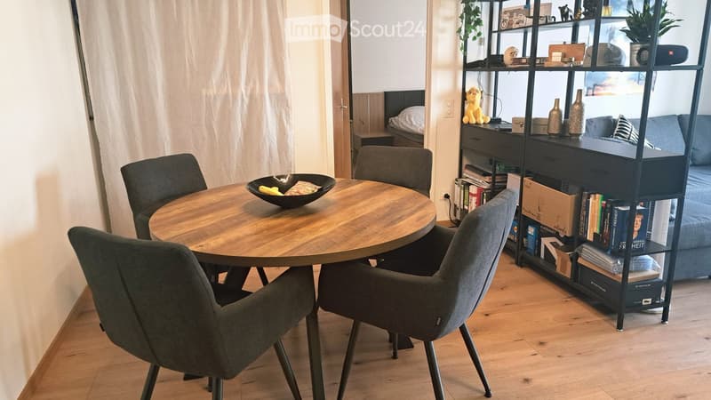 A modern, well-lit dining area with a round wooden table surrounded by four grey chairs. A tall black shelving unit holds books, vases, and decorative items. A small sofa with pillows is visible in the background, along with a plush toy on the shelf.