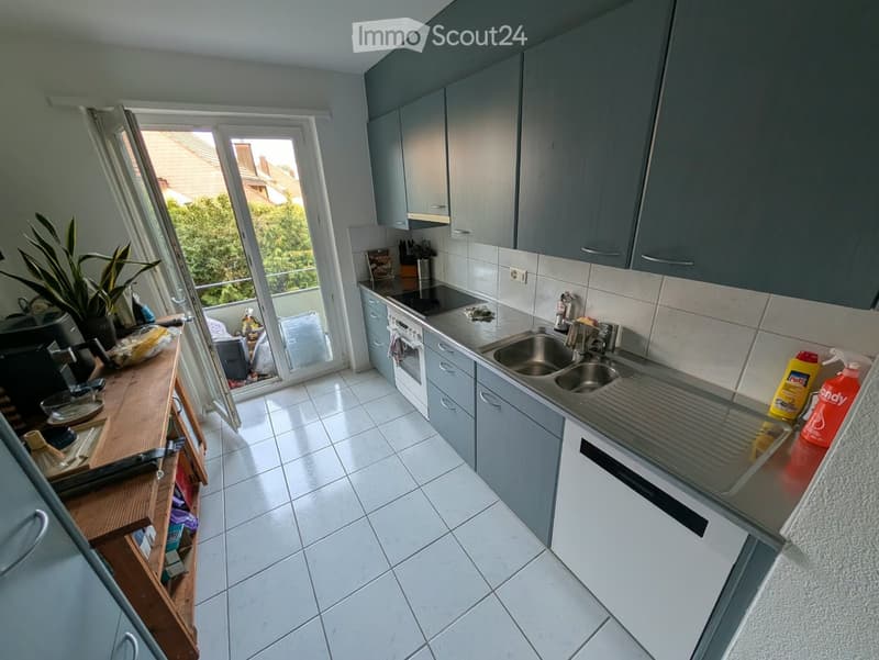 Modern kitchen with grey cabinets, white tiled floor, stainless steel sink, oven, and a view of the balcony and greenery outside.