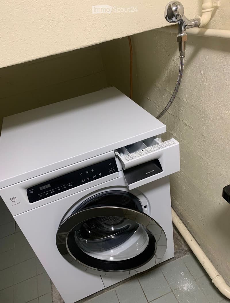 A white washing machine with a control panel and a transparent door is installed in a corner of a room with tiled flooring and beige walls.
