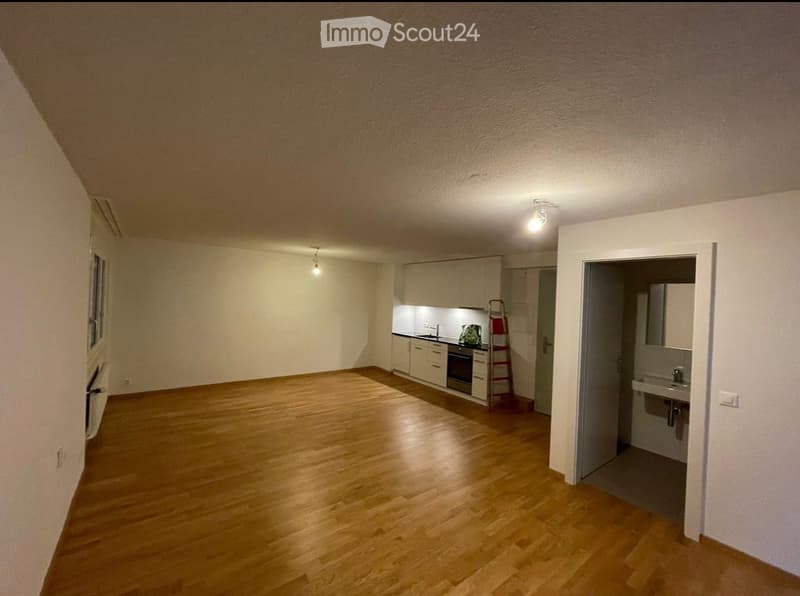 Empty room with wooden flooring, white walls, and a small kitchen area visible in the background. No furniture or personal items are present.