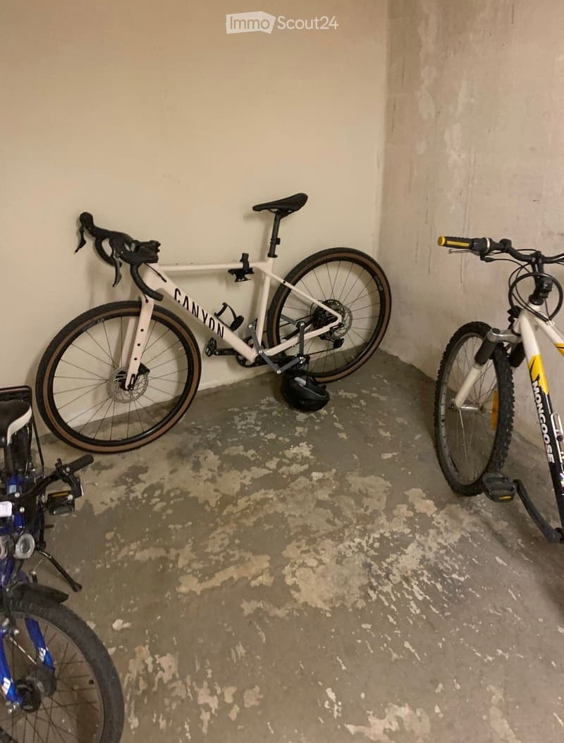 Three bicycles are parked in a small, concrete-walled space. The floor is concrete with some stains, and the walls are plain and light-colored.