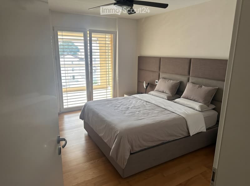 Modern bedroom with a large bed, headboard, nightstand, and lamp. The room features wooden flooring and large windows with blinds.