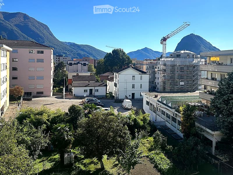 A panoramic view of a town with multiple buildings, including residential and commercial structures, surrounded by greenery and mountains in the background. A construction crane is visible, indicating ongoing development.