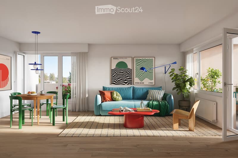 Bright living room with wooden flooring, large windows, a blue sofa with colorful cushions, a red coffee table, a small dining area with a round table and green chairs, and modern lighting fixtures.