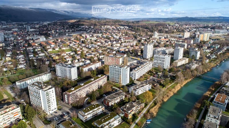 Aerial view of a cityscape with numerous residential and commercial buildings, a river running through the center, and surrounding mountains in the background.
