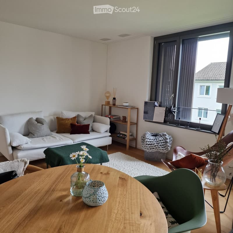 The room features a white couch with cushions, a rug, a wooden table with a glass vase, a wooden cabinet with books and items, and a window with blinds. The floor is wooden, and the walls are white. The room has a cozy and comfortable feel.