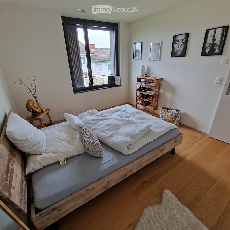A wooden bed with two white pillows, one gray pillow, white sheet and gray blanket. Wooden table with a lamp and a plant on the left. A small wooden shelf filled with shoes, a white bag and a picture frame. Three photo frames on the wall.