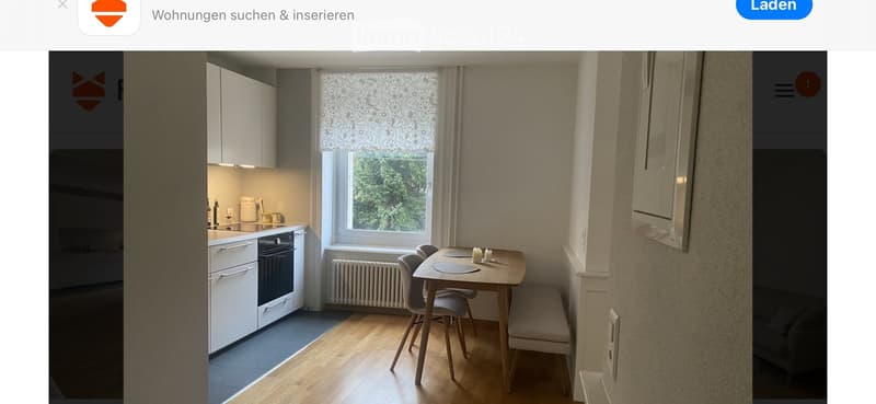Modern kitchen with white cabinets, a sink, an oven, and a small dining area with a table and chairs. The room features wooden flooring and a large window with a lace curtain.