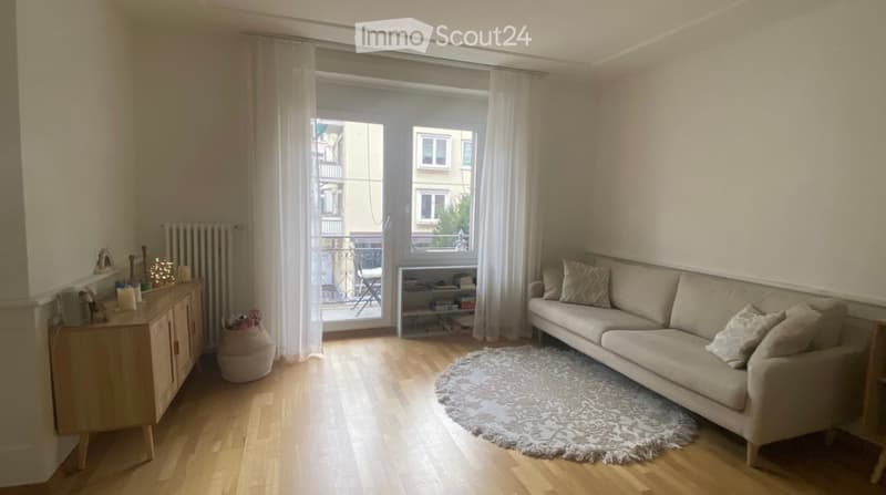 Bright living room with wooden flooring, large window with white curtains, beige sofa with throw pillows, round rug, sideboard with decorative items, and a shelf with books.