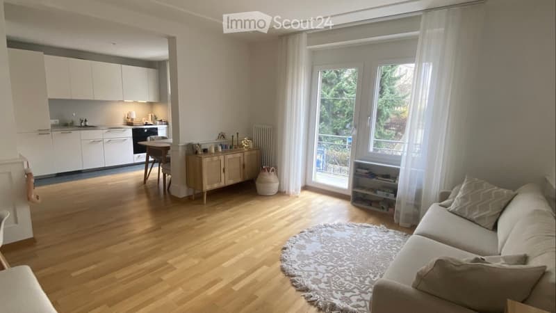 Bright living room with wooden flooring, large windows, a sofa, coffee table, and a small kitchen area with white cabinets and appliances.