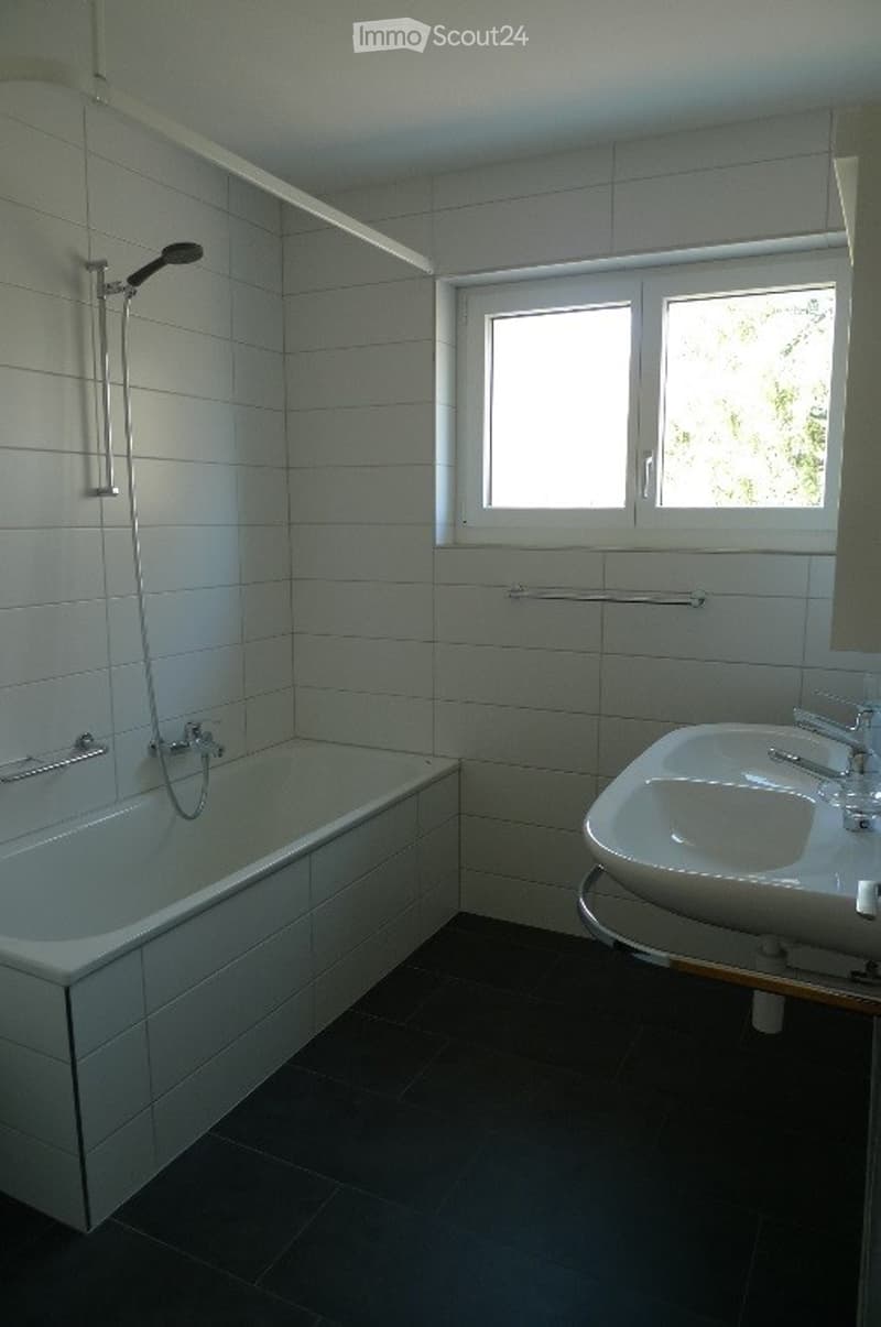 Modern bathroom with white tiled walls and dark tiled floor, featuring a bathtub, a sink with a chrome faucet, and a showerhead with a rail.