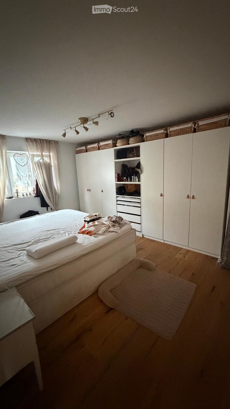 Bedroom with a wooden floor, a bed with white bedding, a wardrobe, and a window with curtains. There are baskets on top of the wardrobe and a small rug on the floor.