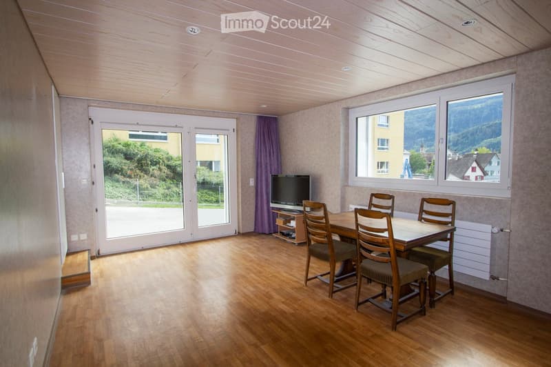 Bright dining area with wooden table and chairs, television on a stand, large windows with scenic mountain view, and sliding glass doors leading outside.