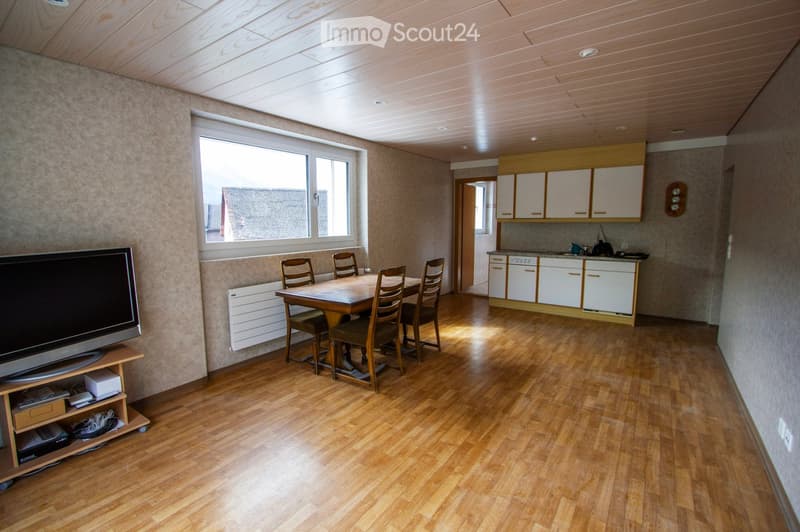 A well-lit room with wooden flooring, a dining table with chairs, a television on a stand, and a kitchen area with cabinets and appliances.