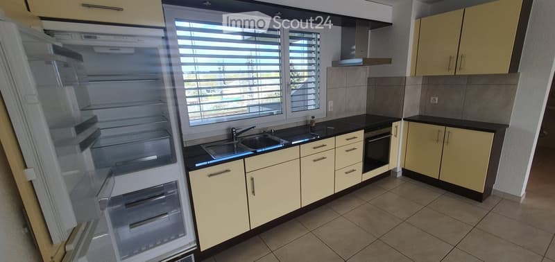 Modern kitchen with light yellow cabinets, black countertops, stainless steel sink, and an open refrigerator. The space features large windows with blinds and a tiled floor.