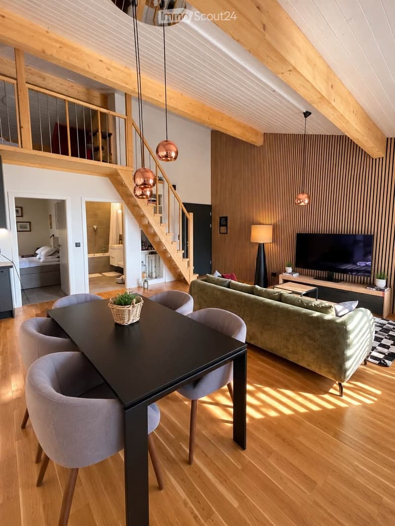 Modern living and dining area with wooden floors, exposed wooden beams, a green sofa, a black dining table with four chairs, a television on a wooden stand, and a staircase leading to an upper level.