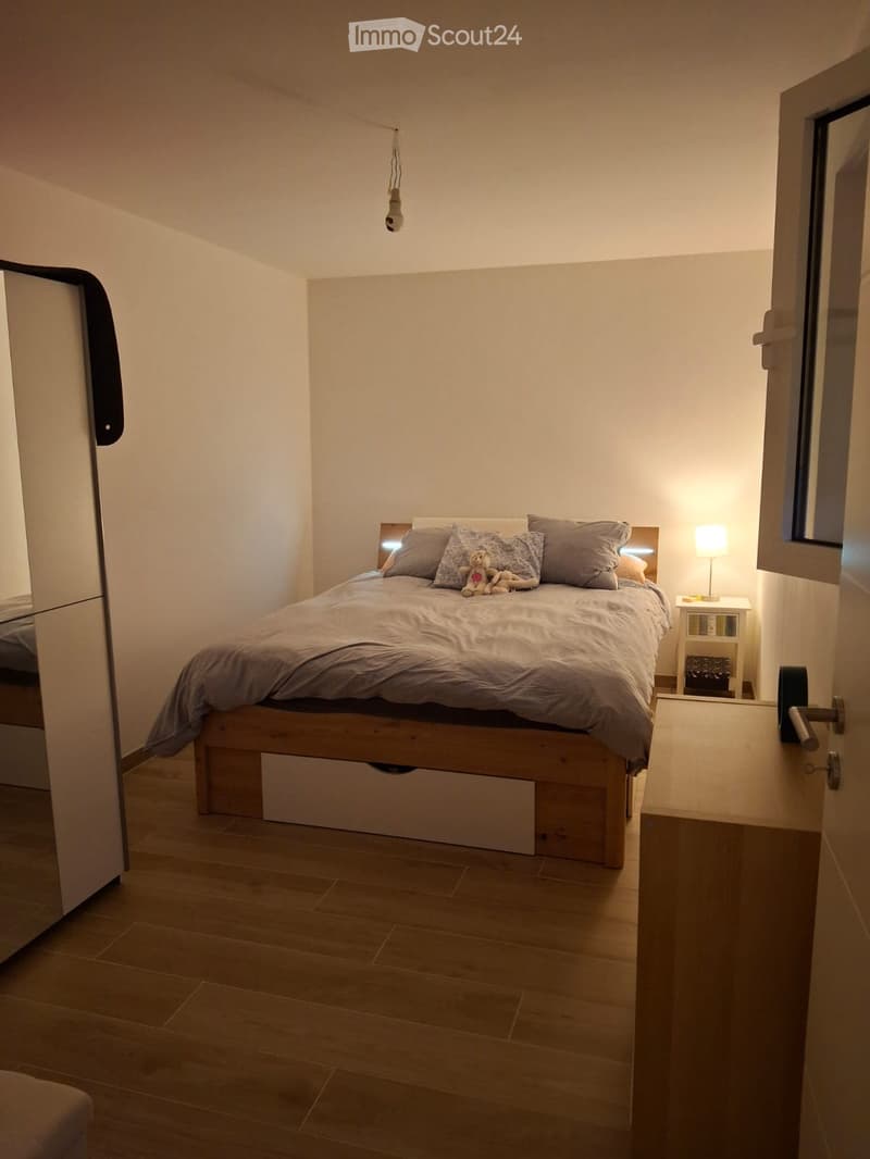 A modern bedroom with a wooden bed, grey bedding, a nightstand with a lamp, and a small toy on the bed.