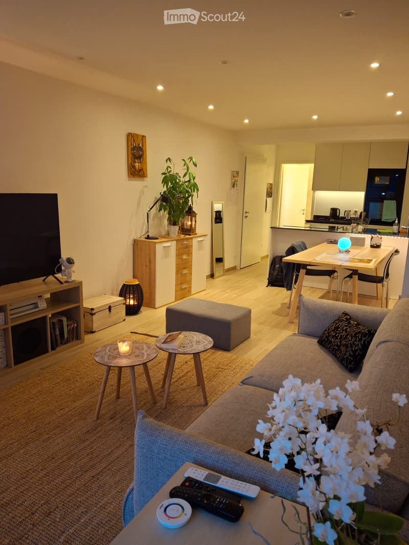 Modern living room with a grey sofa, coffee table, small round tables, a plant, and a kitchen area visible in the background.