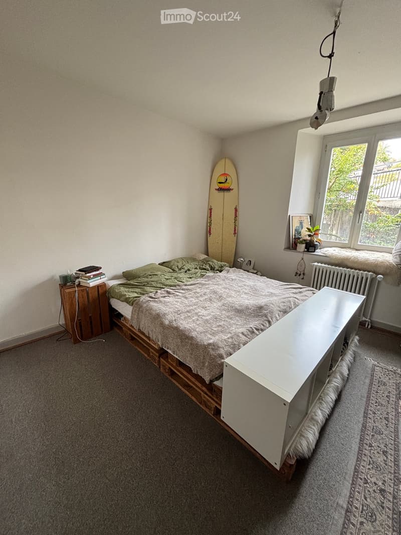 Cozy bedroom with a wooden bed frame, green bedding, nightstand, surfboard leaning against the wall, and a small table near the window.