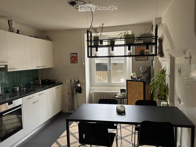 Modern kitchen with white cabinets, black countertops, a dining table with chairs, and a coffee maker on the counter. There is a window with a view, and several plants are placed around the room.