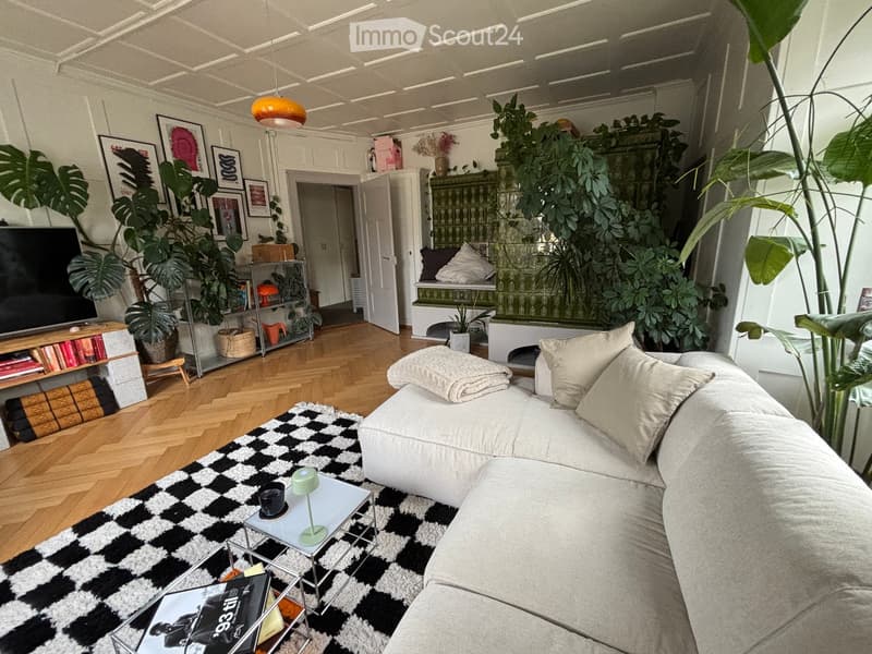Modern living room with a white sofa, coffee table, bookshelves, and large plants. The room features a black and white checkered rug, a television, and several decorative items including picture frames and a side table.