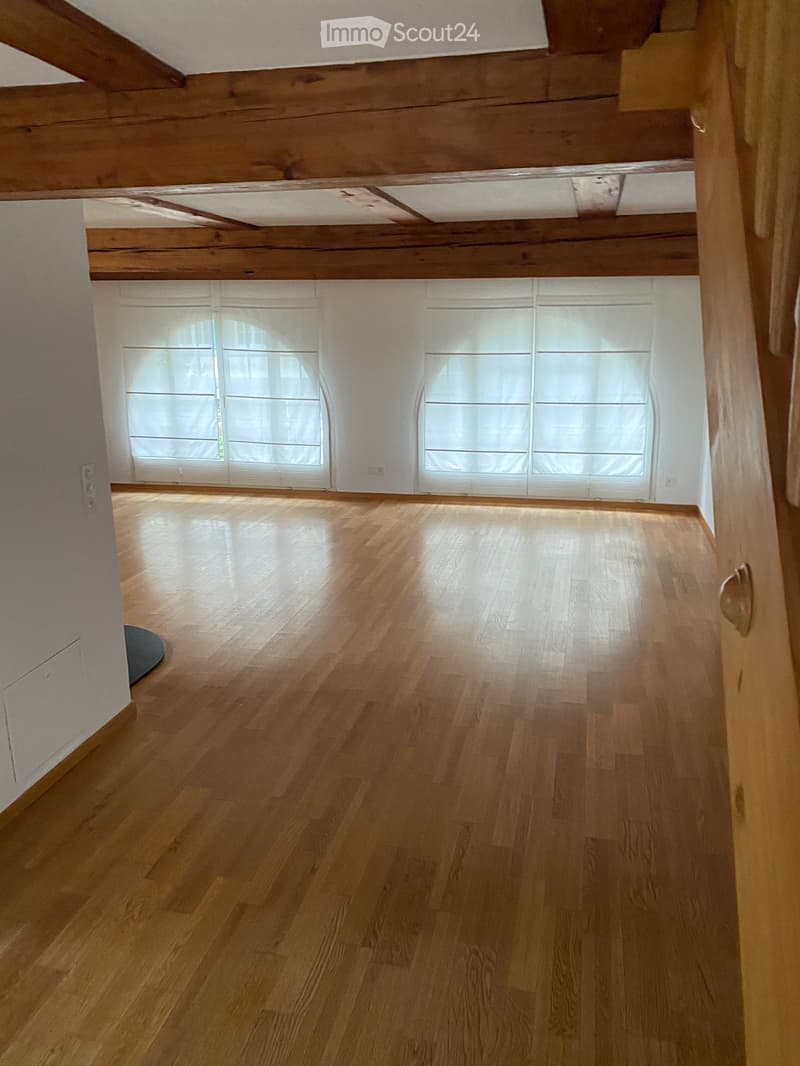 Empty room with wooden flooring, white walls, and large windows with blinds. Exposed wooden beams on the ceiling.