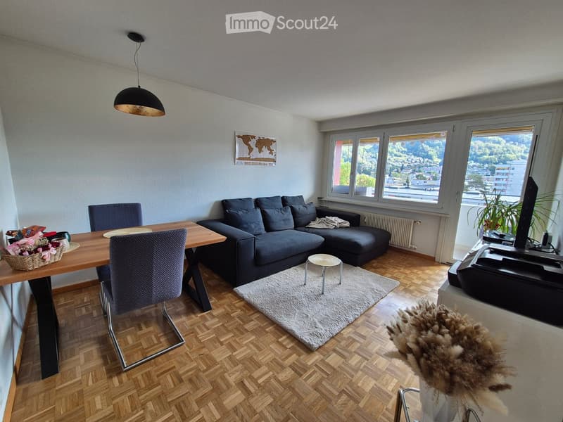 Living room with wooden flooring, large windows offering a scenic view, a grey sectional sofa, a coffee table, a small round stool, a dining table with chairs, a television, and a pendant light.