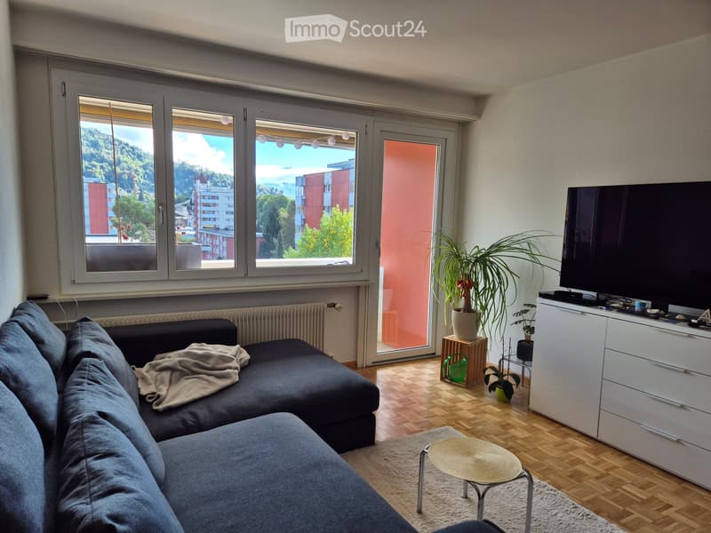 Modern living room with large windows offering a scenic view, a grey sectional sofa, a small round coffee table, a TV on a white cabinet, and several potted plants.