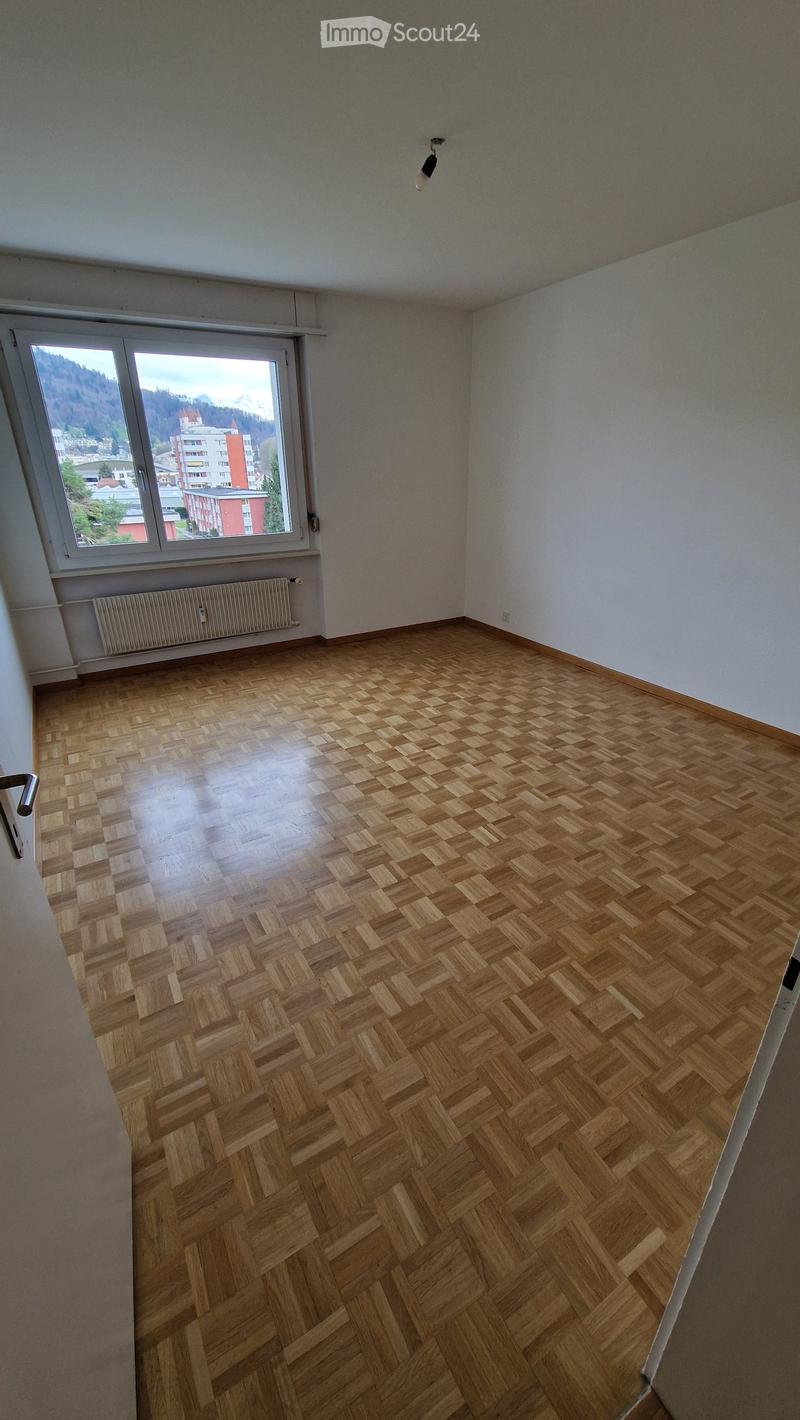 Empty room with wooden herringbone flooring, white walls, large window with view, radiator, and light fixture.