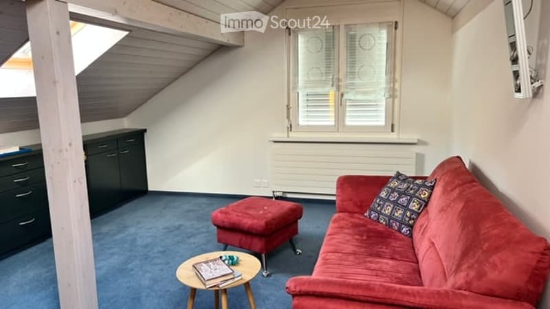 Bright attic living room with slanted ceilings, red sofa, ottoman, small coffee table, and dark wooden cabinet.