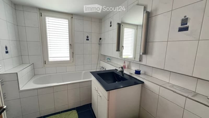Modern bathroom with white tiled walls and floor, a bathtub, a sink with a mirror above it, and a small cabinet below the sink.