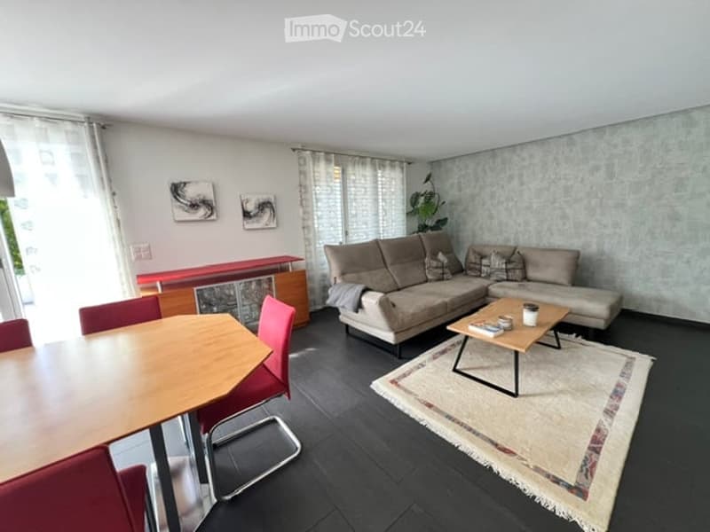 Modern living room with a grey sectional sofa, wooden coffee table, dining table with red chairs, and a patterned rug.
