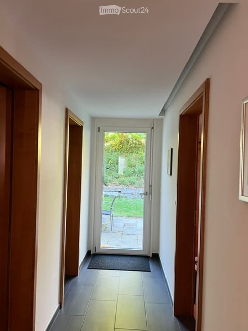A narrow hallway with dark tiled flooring, white walls, and wooden doors on both sides. A glass door at the end leads to a garden area with a bench visible outside.