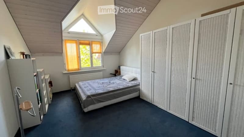 Cozy attic bedroom with slanted wooden ceiling, large dormer window with orange curtains, double bed with patterned bedspread, built-in wardrobes, nightstand, and a mirror on the wall.