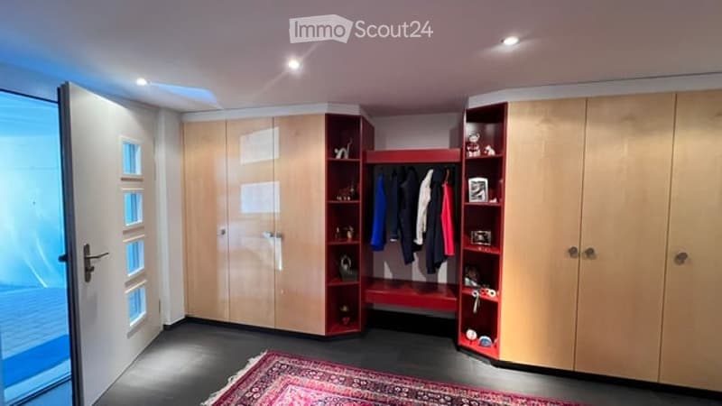 A modern, well-lit walk-in closet with built-in wooden wardrobes, a red shelf unit holding clothing and accessories, and a patterned rug on the floor.