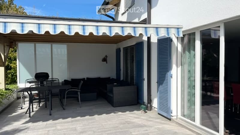 Patio area with a striped awning, outdoor furniture including a sofa, dining table, and chairs, large glass doors leading inside, and blue shutters.