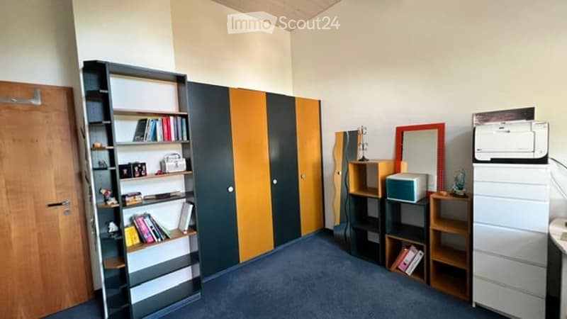 A furnished room with a bookshelf, wardrobe, printer, mirror, table, and chair. The room features a blue carpet and white walls.